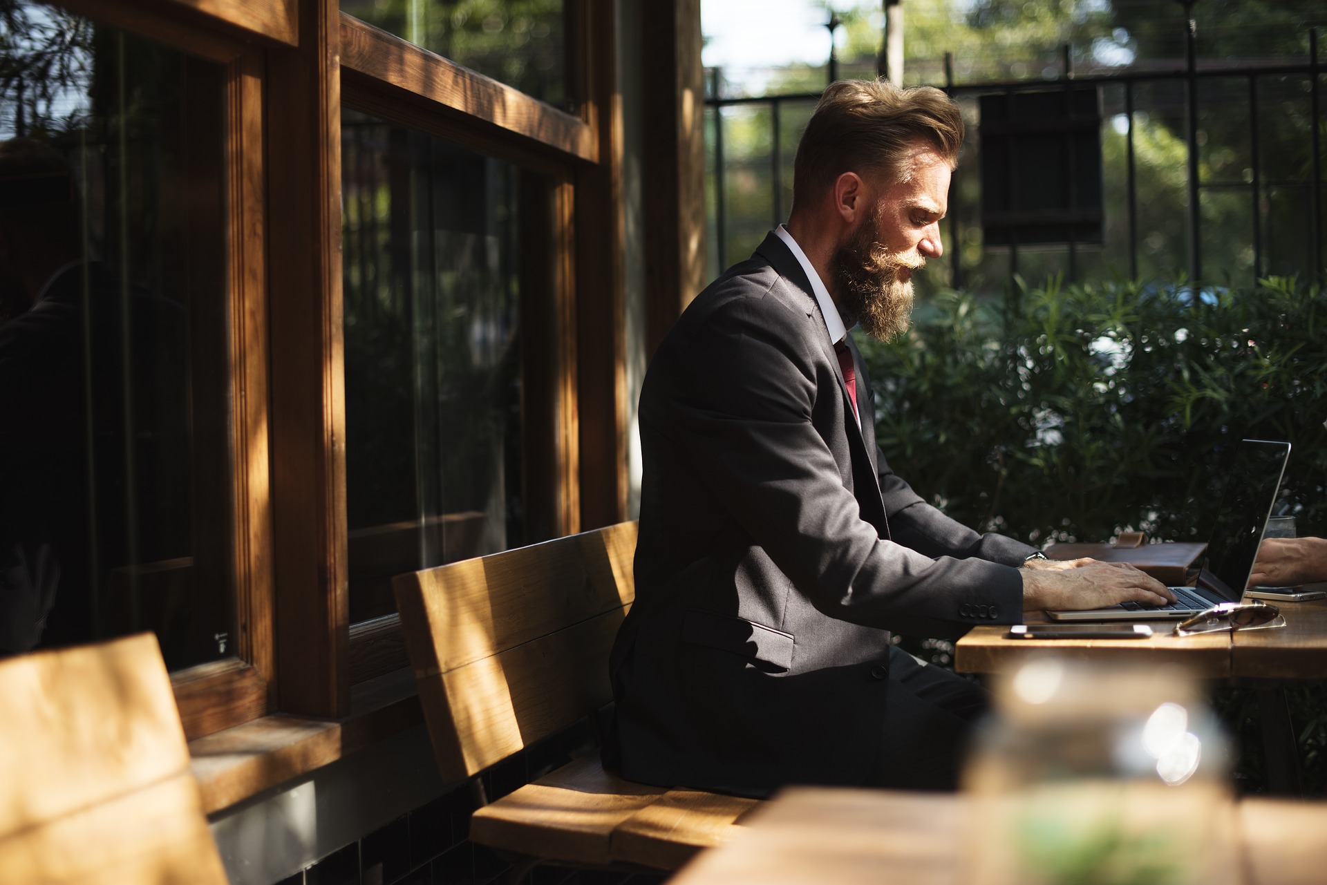 A blond hipster in a suit with a beard uses a laptop in a leafy green coffee shop.